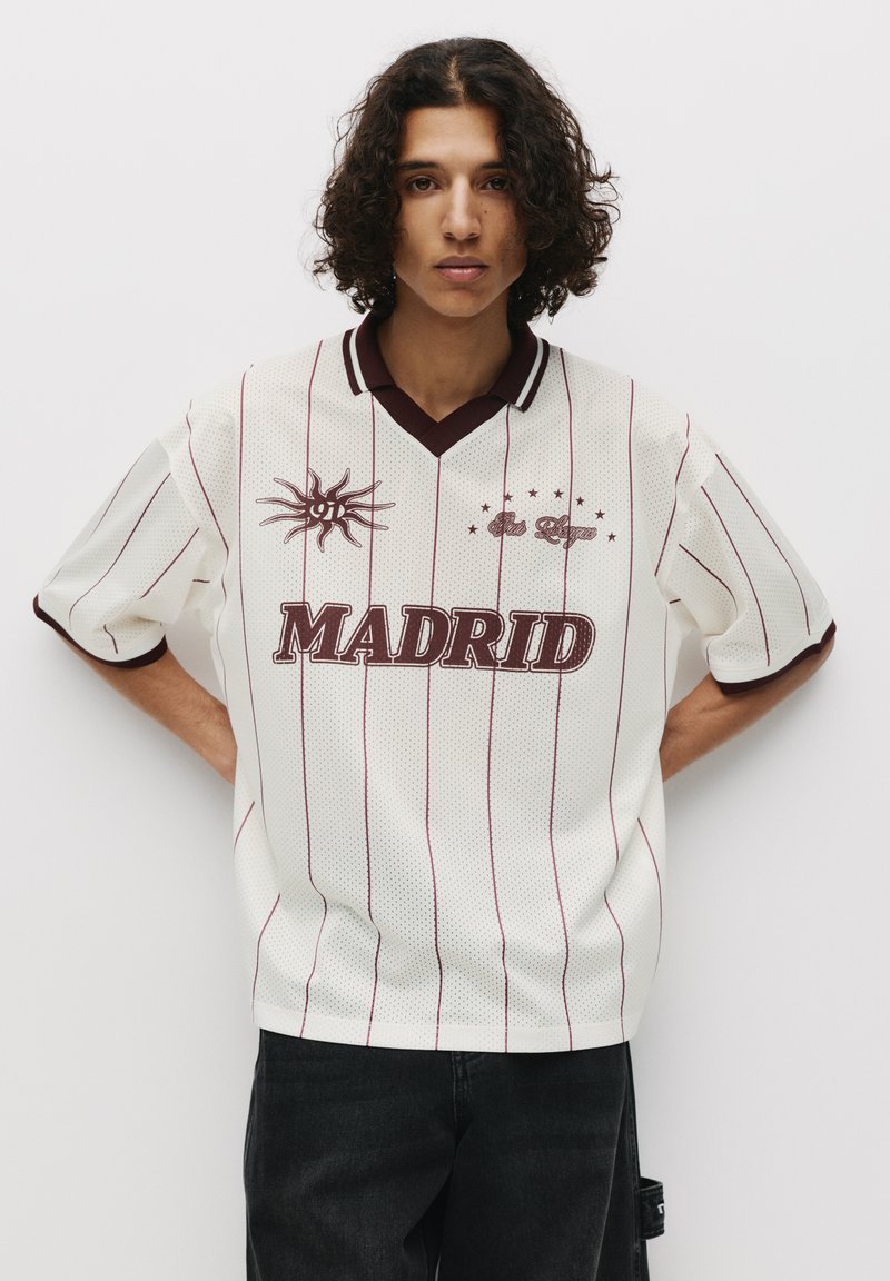 Weißes Mesh-Jersey mit kastanienbraunen Streifen, V-Ausschnitt und dem Text "MADRID". Auf der Vorderseite befindet sich ein Sonnengrafik und Sterne, kurze Ärmel, lässige Passform.