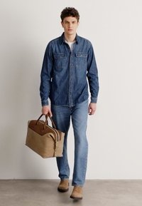 Denim krekls ar pogu aizdari un divām krūšu kabatām, apvienots ar gaiši ziliem džinsiem. Modelis tur tan krāsas ādas somu ar diviem rokturiem.