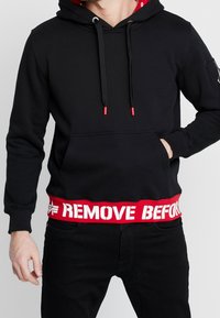 Felpa nera con cappuccio e cordoncino rosso, foderata internamente, dotata di tasca a marsupio e orlo rosso a costine con la scritta bianca "RIMUOVI PRIMA".
