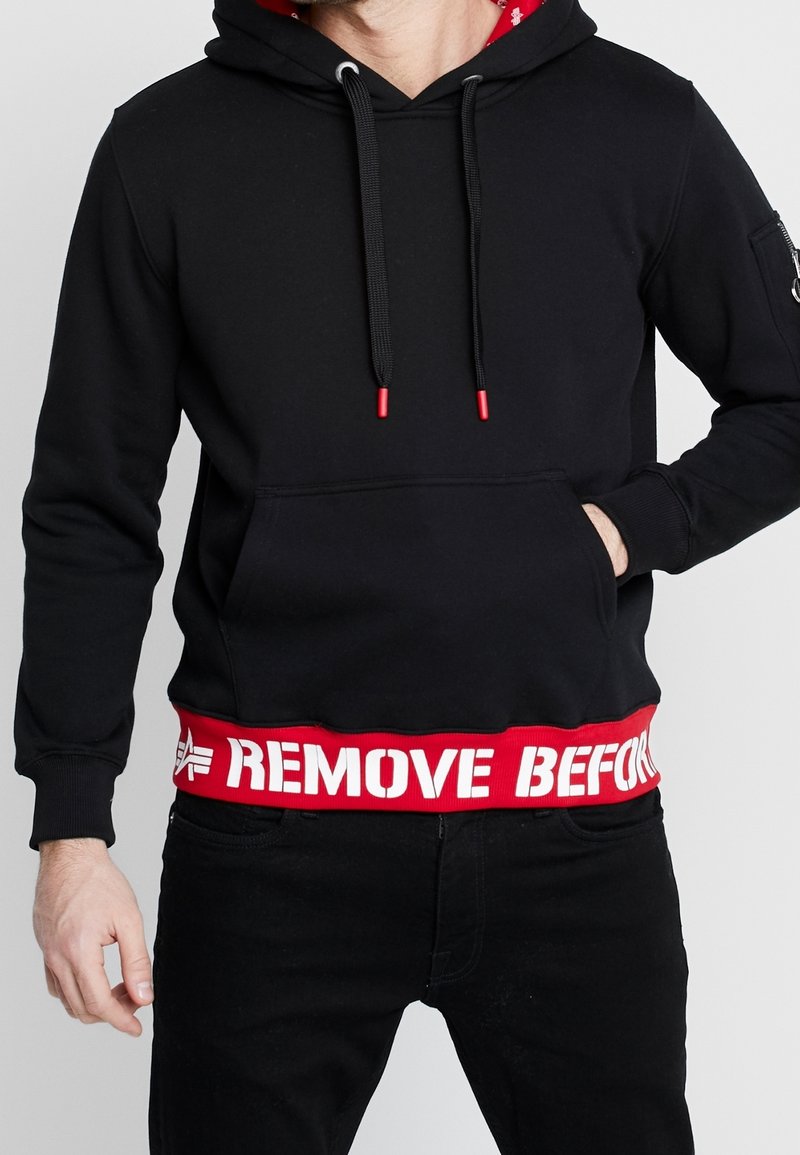 Felpa nera con cappuccio e cordoncino rosso, foderata internamente, dotata di tasca a marsupio e orlo rosso a costine con la scritta bianca "RIMUOVI PRIMA".