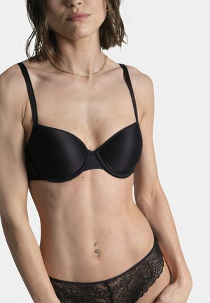 Soutien-gorge rembourré noir avec des détails en dentelle sur les côtés, soutenu par des fines bretelles. Le modèle porte des culottes en dentelle noire assorties.