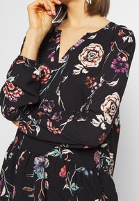Schwarzes Kleid mit langen Ärmeln und floralem Muster, ausgestattet mit einem V-Ausschnitt, elastischen Bündchen und einer gerafften Taille. Bunte Blumen in den Farben Rot, Lila und Türkis.