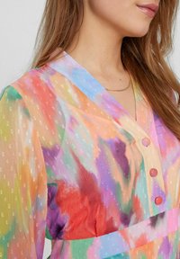 Blouse transparente multicolore avec un col en V, un tissu texturé à petits pois subtils, et trois boutons corail à l'avant.