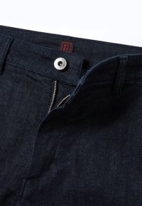 Vaqueros de mezclilla azul oscuro con cierre de cremallera y botón. Se puede ver una etiqueta roja en la cintura, con texto blanco. La tela tiene una textura suave.