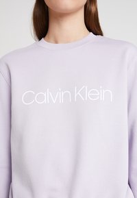 Sudadera de color lila claro hecha de una mezcla suave de algodón. Presenta un cuello redondo y el logo "Calvin Klein" en blanco estampado en el pecho.