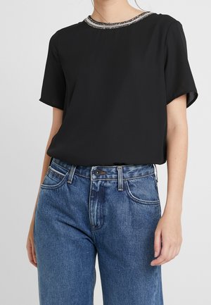 Vrouw draagt een zwart kortermig blouse met een kralenversiering rond de hals, gecombineerd met een high-waist blauwe jeans, staand tegen een effen achtergrond.