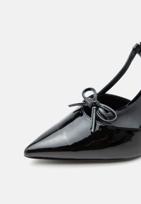 Μαύρο παπούτσι μπαλέτου με μυτερή μύτη από δέρμα με πατέντα, με μικρό φιόγκο μπροστά και λεπτό λουράκι στον αστράγαλο, σε λευκό φόντο.