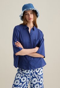 Camisa de botones azul marino de manga corta, combinada con pantalones de patrón floral azul y blanco y un sombrero tipo cubo de mezclilla azul con bordes deshilachados.