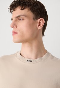 T-shirt beige à col rond avec un col côtelé, comportant une étiquette logo noire au niveau du cou. Texture de tissu lisse, design ajusté.