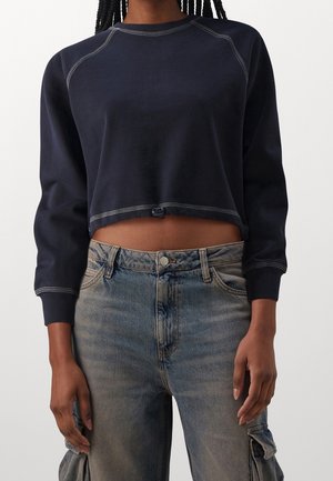 Personne portant un sweat-shirt court à manches longues bleu marine avec des coutures blanches et un jean cargo délavé clair, debout devant un fond uni.
