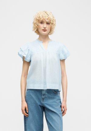 Kvinde med kort krøllet blondt hår iført en lyseblå bluse med flæser og korte ærmer samt højttaljede blå jeans, stående mod en hvid baggrund.