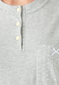 Graues Baumwolloberteil mit runder Halslinie, drei elfenbeinfarbenen Knöpfen und einer aufgesetzten Tasche mit einem weißen Punktmuster und Schleifendetail.