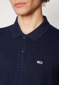 Närbild på en person som bär en marinblå polo t-shirt med två knappar och en liten rektangulär logotyp i vitt, rött och marinblått på bröstet.