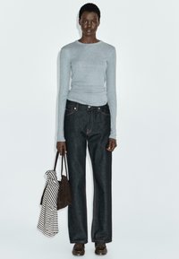 Maglietta grigia a maniche lunghe, jeans a gamba larga blu scuro con cuciture arancioni, borsa marrone e tessuto a righe drappeggiato su un braccio. Design e colori semplici.