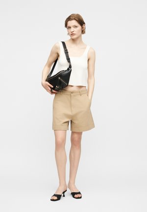 Jauna moteris dėvi baltą be rankovių palaidinę, smėlio spalvos šortus iki kelių, juodus aukštakulnius sandalų ir neša juodą per petį nešamą rankinę.