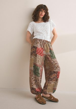 Kvinde med krøllet hår iført hvid kortærmet hæklede top, løse flerfarvede mønstrede bukser og brune åbne sandaler, stående ved en væg.