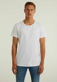 Un tricou alb din bumbac cu guler rotund și mâneci scurte. Prezintă un mic logo pe piept și are o croială lejeră, asortat cu blugi albaștri.