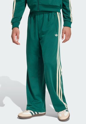 Osoba u zelenim Adidas trenirkama s bež prugama i odgovarajućim tenisicama, stoji ispred jednobojne svijetle pozadine.
