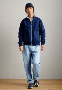 Ναυτικό μπλε μπουφάν bomber με φερμουάρ μπροστά και δύο μπροστινές τσέπες, συνδυασμένο με ανοιχτό μπλε τζιν και μαύρα αθλητικά παπούτσια με λευκές λεπτομέρειες.