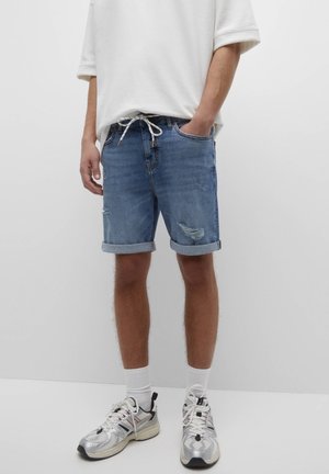 Pantalones cortos de mezclilla en azul con un acabado desgastado, puños enrollados y una cinturilla con cordón. Combinados con calcetines blancos y zapatillas deportivas plateadas.