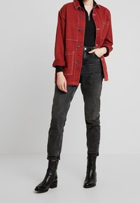 Röd oversizejacka med vit söm, bärs över en svart topp, ihop med figurnära svarta jeans och svarta ankelskostpumpor med låg klack.