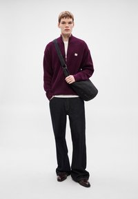 Μπορντό πλεκτό πουλόβερ με φερμουάρ στο γιακά, συνδυασμένο με λευκό μπλουζάκι, σκούρο τζιν παντελόνι και μαύρη τσάντα ώμου. Καφέ παπούτσια ολοκληρώνουν το σύνολο.