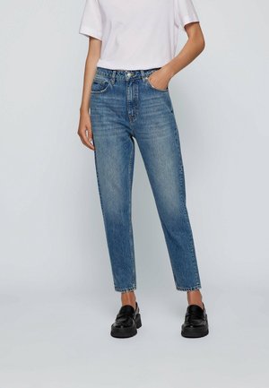 Vrouw draagt high-waist rechte blauwe jeans gecombineerd met een wit T-shirt met korte mouwen en zwarte loafers, staand met één hand in de zak.