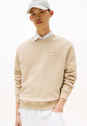 Felpa beige con polsini e orlo a coste, caratterizzata da un piccolo logo sul petto. Indossata sopra una camicia a quadri con colletto.