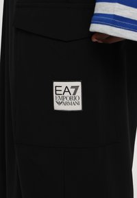 Μαύρο παντελόνι με τετράγωνη λευκή ετικέτα με μαύρο κείμενο που γράφει "EA7 EMPORIO ARMANI." Υφασμάτινη υφή με πλαϊνή τσέπη.