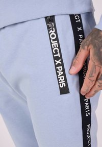 Lichtblauwe joggers met een zwarte zijstreep met daarop de witte tekst "PROJECT X PARIS." Zachte katoenen stof en ritszakken.