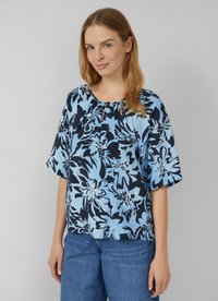Frau, die eine blaue Bluse mit floralem Muster, kurzen Ärmeln und Rundhalsausschnitt trägt, kombiniert mit blauen Jeans, vor einem grauen Hintergrund steht.