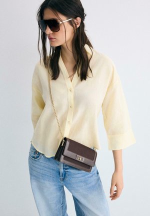 Vrouw met oversized zonnebril, lichtgeel overhemd met knopen, lichtblauwe spijkerbroek en een kleine donkerbruine schoudertas met een gouden ketting.