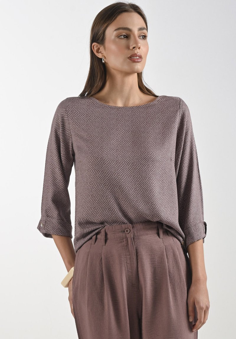 Bruine blouse met patroon en driekwart mouwen, voorzien van een ronde halslijn en opgerolde mouwen, gecombineerd met lichtbruine geplooide broeken.