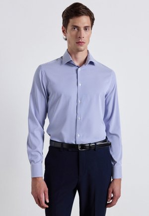 Uomo che indossa una camicia a maniche lunghe a righe azzurro chiaro infilata in pantaloni scuri con cintura nera, in piedi davanti a uno sfondo semplice.