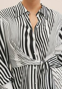 Chemise à rayures noires et blanches en tissu lisse. Elle possède un col, une fermeture par boutons, et un détail de taille nouée avec des motifs de rayures variés.