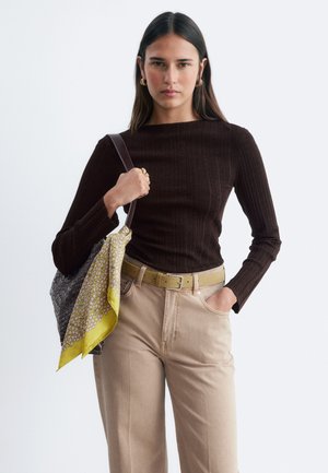 Femme portant un haut côtelé marron foncé à manches longues, un pantalon beige avec ceinture, tenant un sac tressé avec une écharpe jaune à motifs.