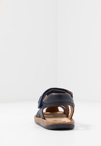 Granatowy skórzany sandał z odkrytą piętą, regulowanym paskiem i brązową korkową podeszwą. Posiada niebieski akcent w okolicy pięty oraz przeszycia dekoracyjne.