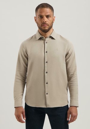 Man met kort haar en een baard, draag een beige overhemd met knopen en donkere broek, staand tegen een effen lichte achtergrond.