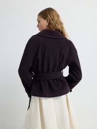 Giacca viola, cintrata con colletto e dettaglio arricciato sulla schiena, abbinata a una gonna bianca fluente; texture liscia e design strutturato.