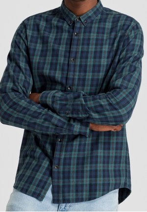 Mężczyzna w zielono-granatowej koszuli w kratę z długim rękawem i zapiętej na guziki, ze skrzyżowanymi ramionami, w połączeniu z jasnoniebieskimi dżinsami.