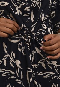 Granatowa sukienka z białym wzorem liści, z zapięciem na guziki z przodu oraz detalem w postaci związania w talii. Gładki materiał o luźnym kroju i długich rękawach.