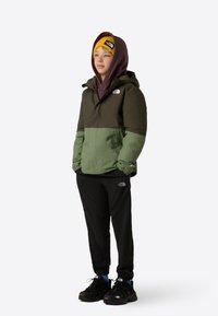 Jachetă rezistentă la apă, de culoare verde închis și măslinie, cu glugă și manșete elastice, purtată peste un hanorac burgund și pantaloni sport negri.