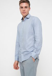 Sininen ja valkoinen ruudullinen nappikauluspaita, jossa on kaulus, pitkät hihat ja hieman kapeneva leikkaus, yhdistettynä vaaleanharmaisiin housuihin.