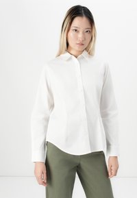 Young woman with straight ombre hair wearing a white button-up shirt and olive green pants, standing against a plain light background.