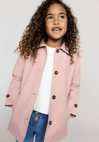 Vaaleanpunainen vedenkestävä takki ruskeilla napilla, rento istuvuus ja avoin kaulus, jota on käytetty valkoisen neulotun neuleen ja sinisten farkkujen päällä.