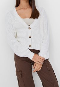 Bílý pletený kardigan s hlubokým výstřihem do V, zapínáním na knoflíky, žebrovanými rukávy a měkkou, texturovanou tkaninou, nošený přes světlý top.