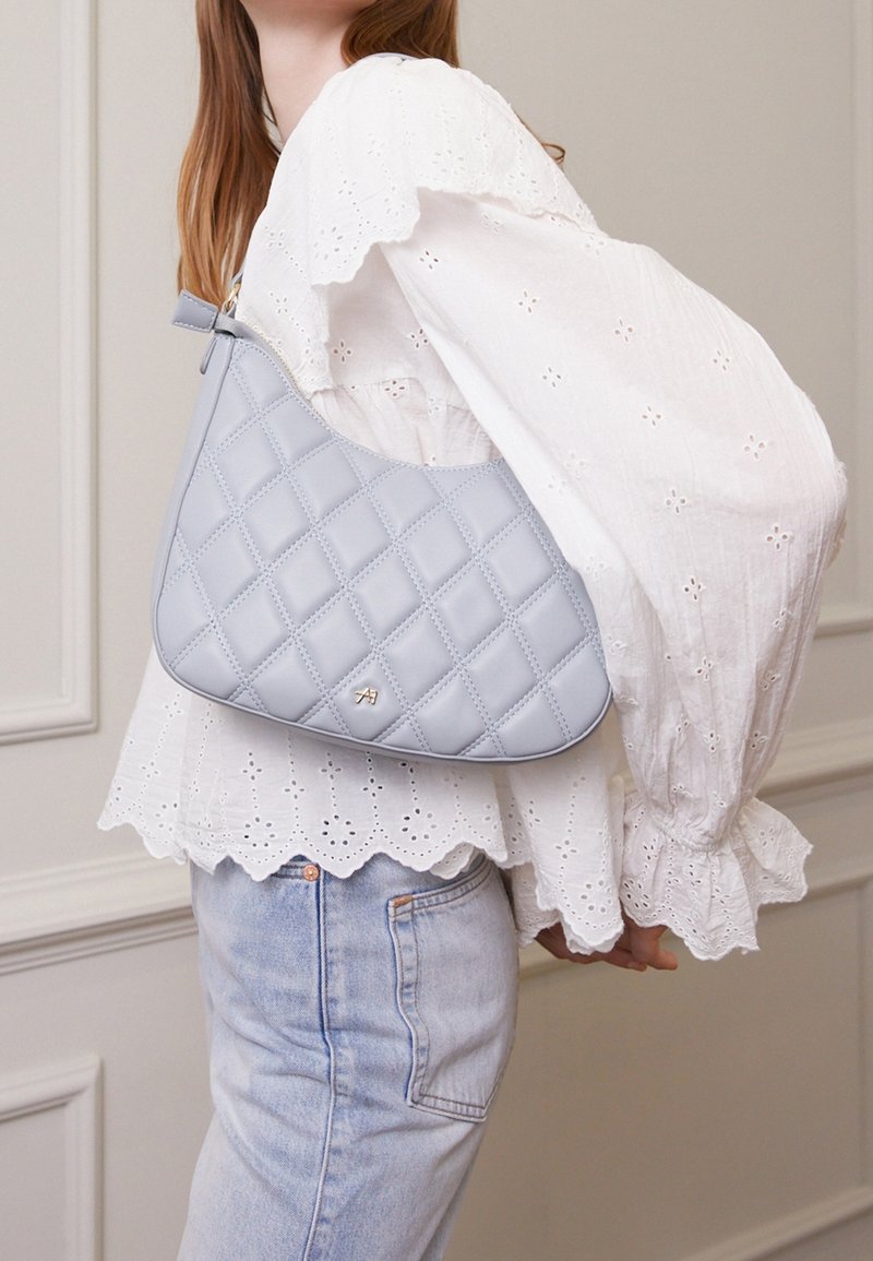 Vaaleansininen tikkikassi, jossa on kaareva muoto ja sileä pinta. Varustettu kultaisilla yksityiskohdilla ja sitä kannetaan valkoisen broderie anglaise -paidatoppauksen päällä.