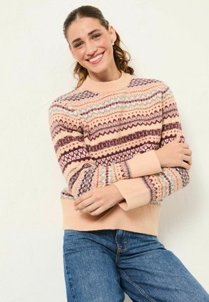 Knit sweater featuring a light pink base with multicolored geometric patterns and ribbed cuffs. Styled with blue denim jeans.
