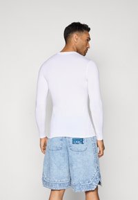 Hvid langærmet skjorte parret med lyseblå denimshorts, der har en dekorativ kant og logo på baglommen. Glat overflade generelt.