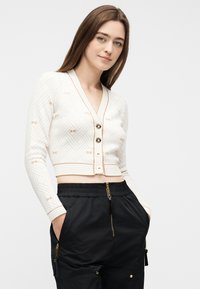 Νεαρή γυναίκα που φοράει ένα λευκό cropped ζακετάκι με σχέδια και χρυσά κουμπιά, καθώς και μαύρο παντελόνι με φερμουάρ στις τσέπες, ποζάροντας με τα χέρια στις τσέπες.